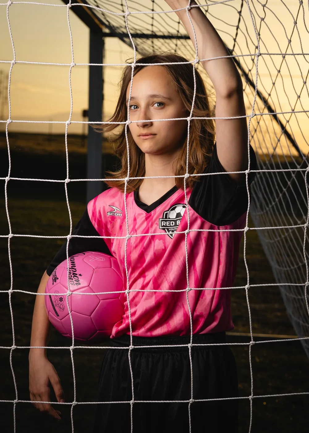 Soccer player in a pink kit looking through the goal net at sunset